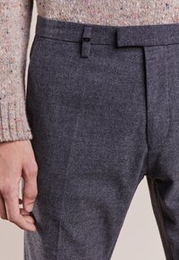 Pantaloni grigi in tessuto intrecciato con una finitura testurizzata, caratterizzati da una frontale piatta, passanti per la cintura e una chiusura a gancio, abbinati a un maglione in maglia multicolore.