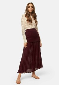 Lace long-sleeve top in ivory, paired with a burgundy pleated maxi skirt and light pink textured flats. Elegant design with soft materials.