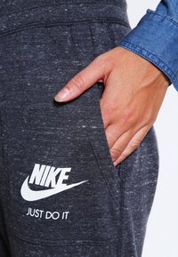 Nike tepláky v tmavě šedém melírovaném materiálu s hladkou texturou. Na levé kapse se nachází bílé logo a text "JUST DO IT".