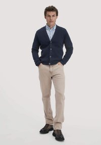 Marineblauwe button-up cardigan over een lichtblauw shirt, beige broek en donkere schoenen. De cardigan heeft een V-hals en geribde manchetten.