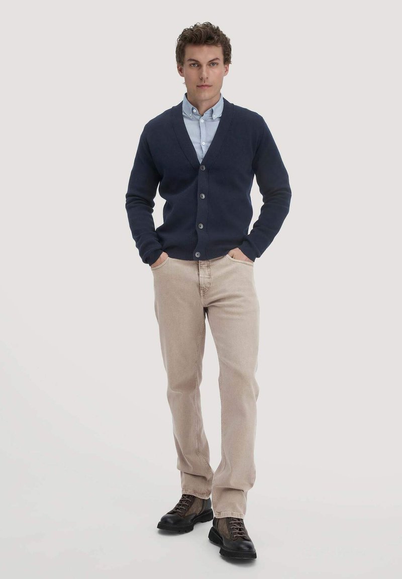 Marineblauwe button-up cardigan over een lichtblauw shirt, beige broek en donkere schoenen. De cardigan heeft een V-hals en geribde manchetten.