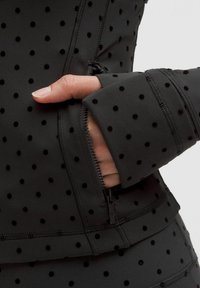 Chaqueta atlética negra con una superficie texturizada que presenta pequeños puntos negros. La vista de cerca muestra un bolsillo con cremallera y detalles en los puños.