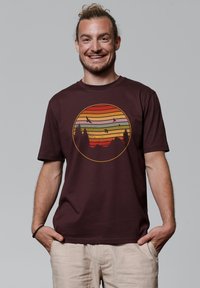 Bordeaux kleurige t-shirt met korte mouwen, met een cirkelvormige afbeelding van een zonsondergang met gelaagde strepen, bergen en vliegende vogels. Gladde katoentextuur.