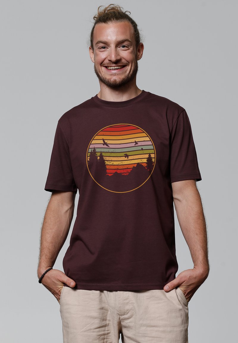Bordeaux kleurige t-shirt met korte mouwen, met een cirkelvormige afbeelding van een zonsondergang met gelaagde strepen, bergen en vliegende vogels. Gladde katoentextuur.