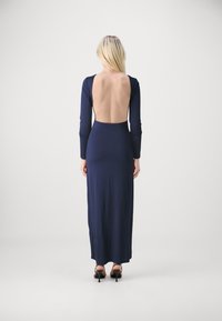 Navy long-sleeve dress with a low back, made of smooth fabric. Fitted silhouette, floor-length, simple design, worn with heeled sandals.
