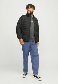 Schwarze gesteppte Daunenjacke mit hohem Kragen, getragen über einem grauen Sweatshirt, kombiniert mit blauen Jeans und schwarzen Sneakers; lockere Passform, sichtbare Nähte.