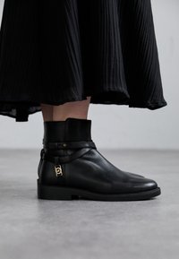Bottines en cuir noir avec un bout arrondi, semelle plate et une sangle décorative. Accents en matériau doré près de la cheville. Jupe en tissu texturé en arrière-plan.