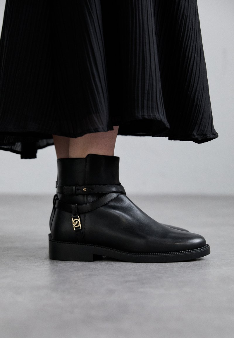 Bottines en cuir noir avec un bout arrondi, semelle plate et une sangle décorative. Accents en matériau doré près de la cheville. Jupe en tissu texturé en arrière-plan.