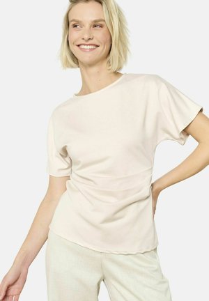 Haut beige clair à manches courtes en tissu doux avec un col rond et un design drapé subtil, associé à un pantalon texturé clair.