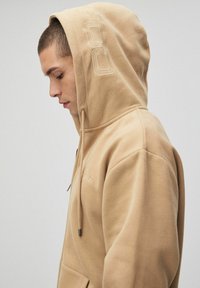 Beżowa bluza z kapturem wykonana z miękkiej mieszanki bawełny, z kapturem na sznurku, przednią kieszenią oraz subtelnym wyszywanym logo na klatce piersiowej i rękawie.