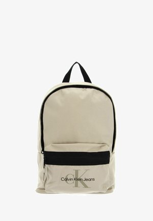 Mochila beige de tela con un bolsillo con cremallera negro. Cuenta con un asa superior negra y el logo de Calvin Klein Jeans en verde oscuro en la parte frontal.