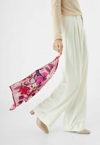 Étole en soie avec des motifs floraux roses et des bordures rouges, tenue dans une main. Une paire de pantalons larges blancs et des chaussures à talons carrés beiges sont visibles.