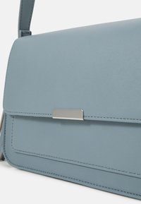 Borsa in pelle blu chiaro con una fibbia rettangolare argento e una sola tracolla, mostrata in primo piano su uno sfondo bianco.