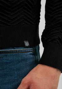 Suéter negro con textura y un patrón en zigzag, puños acanalados y una pequeña etiqueta de logo en el dobladillo, combinado con jeans de mezclilla azules.