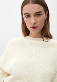 Hvid ribbet striktrøje med høj hals. Blød tekstur og afslappet pasform, vist på en model med glat hår foran en neutral baggrund.