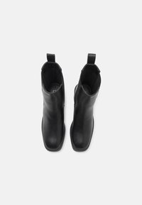 Paire de bottines en cuir noir avec bouts carrés et languettes sur fond blanc, vues de dessus.