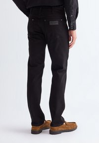 Persoană purtând blugi negri Wrangler și mocasini din piele întoarsă maro, stând cu spatele către privitor pe un fundal simplu.