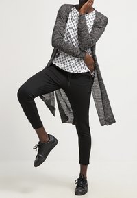 Larga chaqueta gris sobre una camisa blanca con un patrón negro, combinada con pantalones negros ajustados y zapatos negros de cordones.