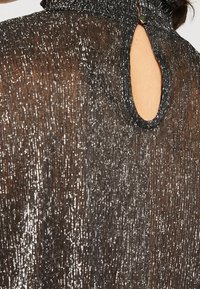 Haut en tissu noir transparent avec des détails en paillettes argentées et une petite ouverture en forme de trou de serrure au niveau du col, fermée par un bouton.