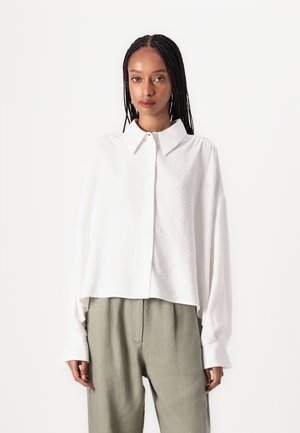 Chemise blanche à manches longues avec un col et une coupe décontractée, présentant une texture de tissu doux et un ourlet court. Associée à un pantalon vert clair.