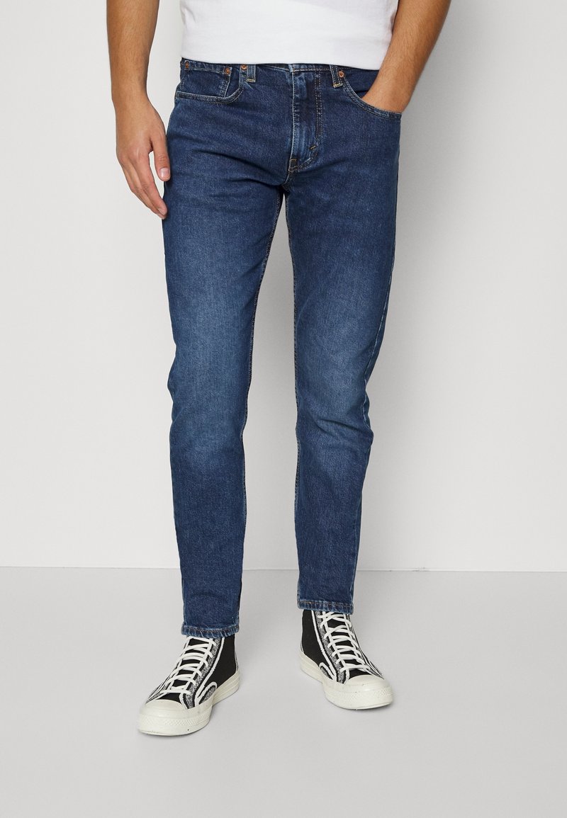 Homme portant un jean slim bleu foncé, main dans la poche, et des baskets montantes noires avec des lacets et semelles blanches, debout sur un sol gris.