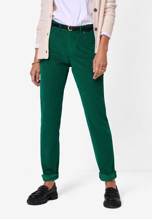 Femme portant un pantalon en velours côtelé vert retroussé aux chevilles, ceinture noire, cardigan beige, chemise blanche, mocassins noirs, debout sur fond blanc.