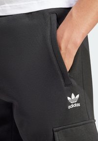 Svarta träningsbyxor i texturerat tyg med sidofickor; har en vit Adidas-logotyp på vänstra fickområdet.