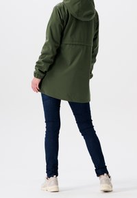 Grön vattentät jacka med huva, avslappnad passform, elastiska manschetter och en mjuk textur, i kombination med mörkblå slim jeans och ljusa sneakers.