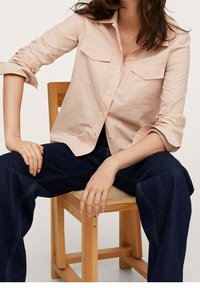 Chemise boutonnière rose clair avec deux poches poitrine, manches retroussées, portée avec un pantalon large bleu marine, assis sur une chaise en bois.