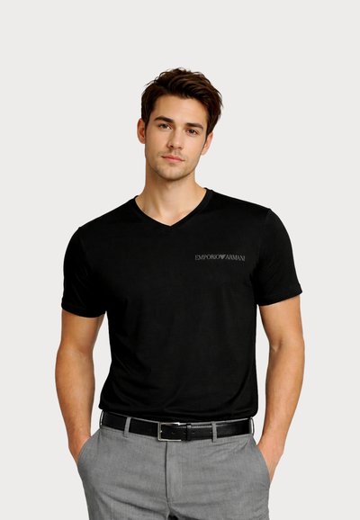 Νεαρός άνδρας που φοράει μαύρο T-shirt Emporio Armani με V-λαιμόκοψη, φορεμένο μέσα σε γκρι παντελόνια με μαύρη ζώνη, τα χέρια στις τσέπες, ουδέτερο φόντο.