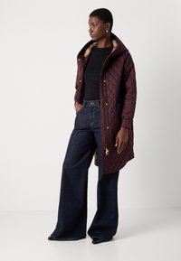 Gequilted kastanjebruine jas met een capuchon en gouden details, gedragen over een zwart shirt, gecombineerd met wijde, donkere denim jeans.