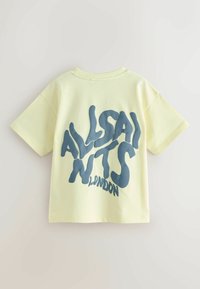 Šviesiai geltona trumpomis rankovėmis marškinėliai, ant nugaros ryškiai matoma mėlyna, skystos formos grafika su užrašu "ALLSAINTS LONDON".