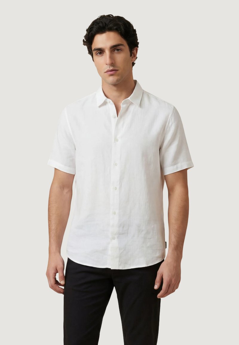 Homme portant une chemise blanche à manches courtes boutonnée et un pantalon noir, debout devant un fond gris clair uni.