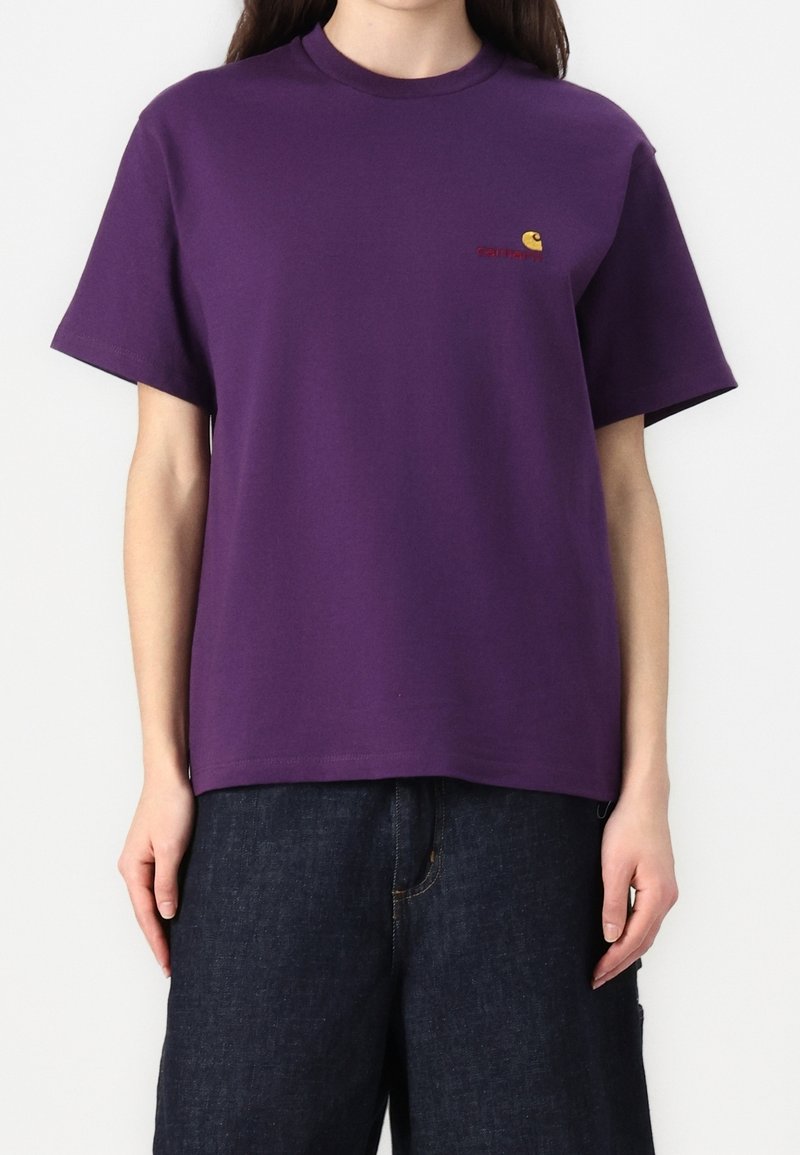 Een persoon draagt een paarse T-shirt met korte mouwen met een klein geel en rood logo op de linkerborst en donkerblauwe spijkerbroek.