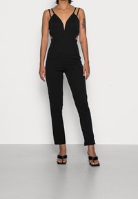Czarny kombinezon z głębokim dekoltem w kształcie litery V, cienkimi ramiączkami i wycięciami po bokach. Dopasowany fason z prostymi nogawkami i minimalistycznym designem.