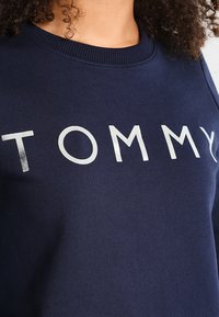 Lockig person med en marinblå sweatshirt där ordet "TOMMY" är tryckt i stora vita bokstäver på bröstet.