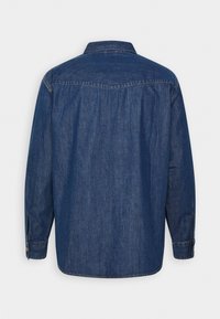 Chemise en denim bleu foncé à manches longues avec poignets boutonnés vue de dos, présentant une couture de renfort au-dessus du dos.