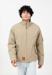 Beżowa, wodoodporna kurtka z wysokim kołnierzem, zapinana na zamek, z bocznymi kieszeniami i pomarańczową metką u dołu. Teksturowana tkanina i dopasowany krój.