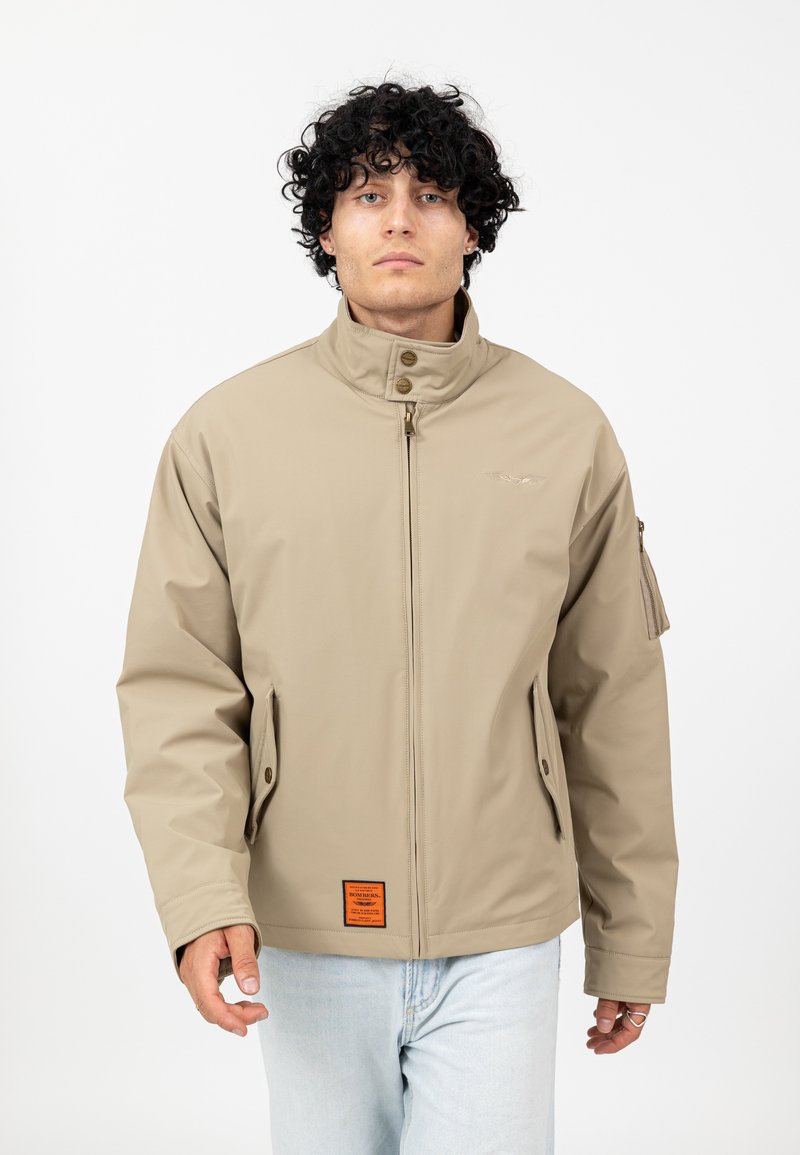 Beżowa, wodoodporna kurtka z wysokim kołnierzem, zapinana na zamek, z bocznymi kieszeniami i pomarańczową metką u dołu. Teksturowana tkanina i dopasowany krój.