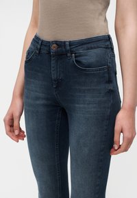 Femme portant un jean bleu foncé ajusté et un haut beige sans manches, debout les bras détendus le long du corps.