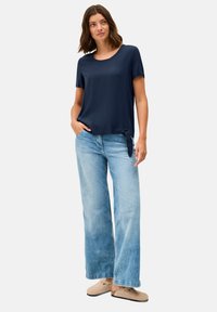 T-shirt navy à manches courtes avec un col rond et un détail de nœud sur le côté, associé à un jean large bleu clair et des chaussures beiges à enfiler.