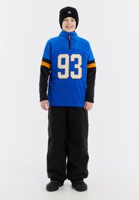 Niebieski sweter z półzamkiem, z dużym numerem 93 w białym kolorze, czarnymi rękawami z pomarańczowymi paskami, w zestawie z czarnymi spodniami narciarskimi i butami.