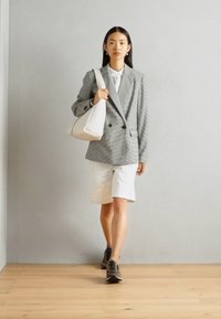Grijs geruit dubbelknopen blazer, wit shirt, witte shorts en donkere sneakers. Model draagt een gebogen witte handtas en staat op een houten vloer.