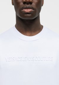 Zbliżenie osoby noszącej białą koszulkę z okrągłym dekoltem, na której na piersi znajduje się wytłoczony napis „VERSACE JEANS COUTURE”.