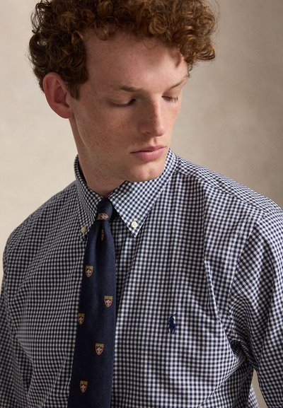 Jeune homme aux cheveux roux bouclés, portant une chemise à carreaux bleue et blanche et une cravate bleu marine avec de petits écussons, regardant vers le bas et sur le côté.