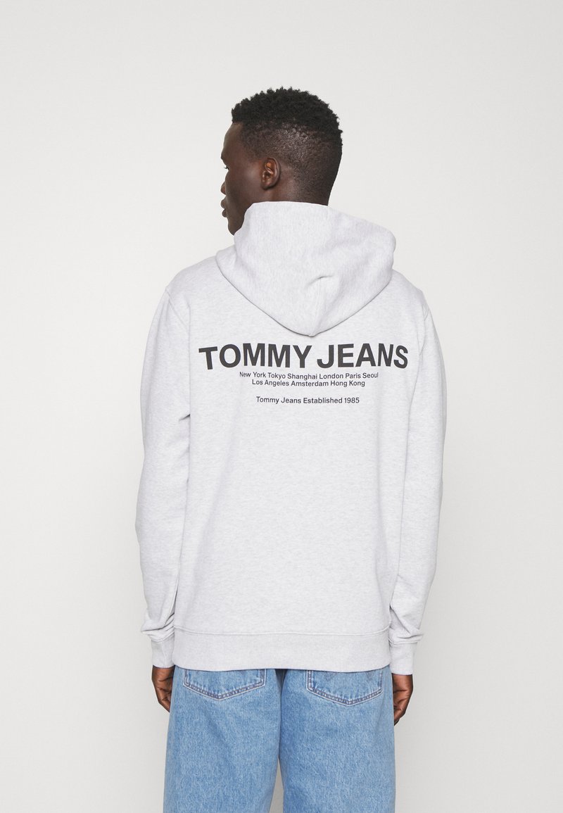 Sudadera gris con capucha. Presenta el logotipo "TOMMY JEANS" en la parte posterior en negro, junto con nombres de ciudades y la fecha de establecimiento. Ajuste casual.