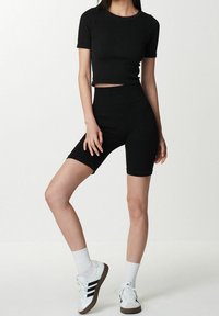 Femme portant un t-shirt noir ajusté et court avec un short de vélo noir taille haute, des chaussettes blanches et des baskets blanches avec des rayures noires.