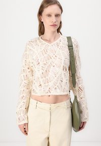 Häkelpullover in Off-White, mit offenen Webstrukturen und langen Ärmeln. Kombiniert mit beigen Hosen und einer grünen Umhängetasche.