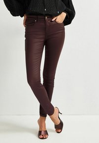 Jeans skinny în nuanță burgundy, dintr-un material neted și elastic, cu buzunare frontale și un accent dintr-un buton metalic, asortați cu sandale cu vârf deschis, de culoare maro.