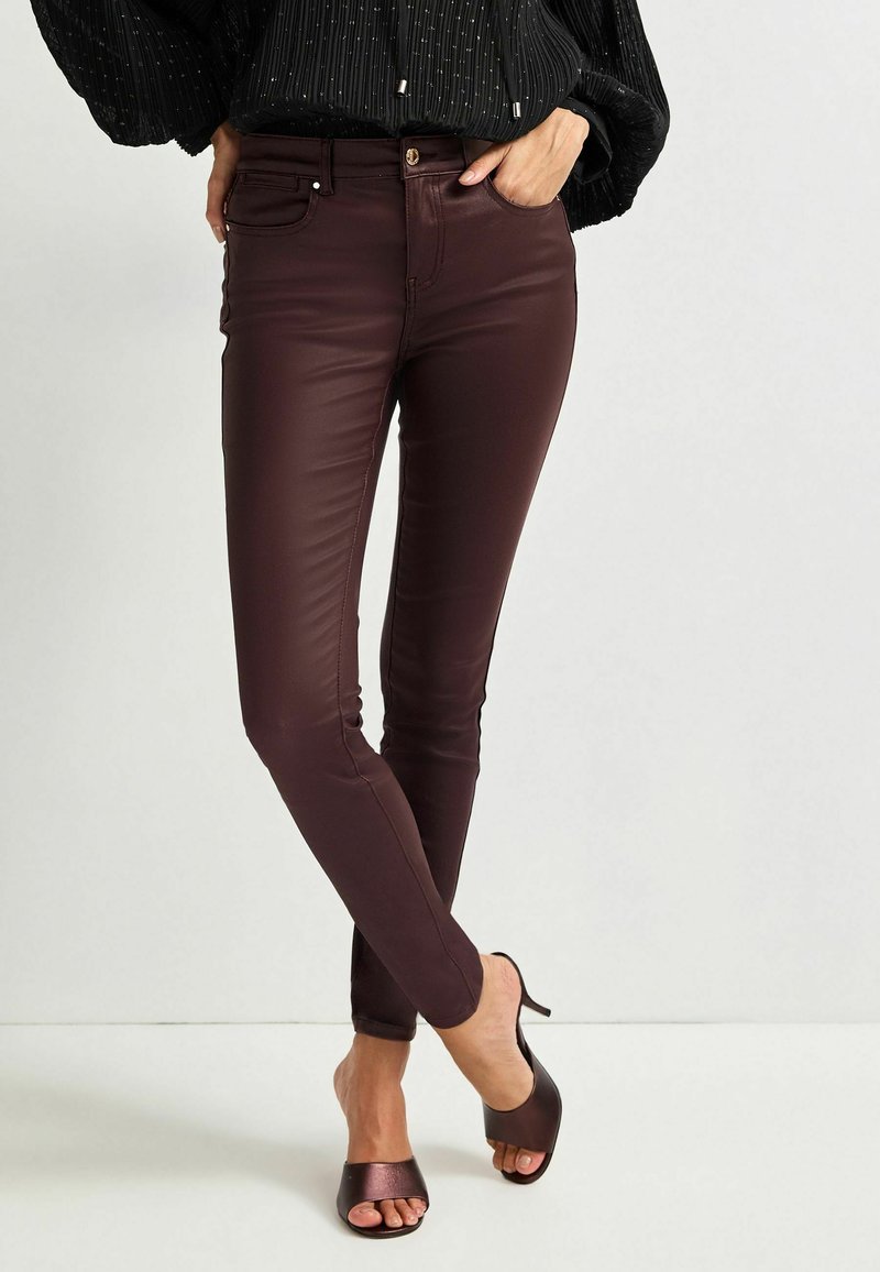 Jeans skinny în nuanță burgundy, dintr-un material neted și elastic, cu buzunare frontale și un accent dintr-un buton metalic, asortați cu sandale cu vârf deschis, de culoare maro.